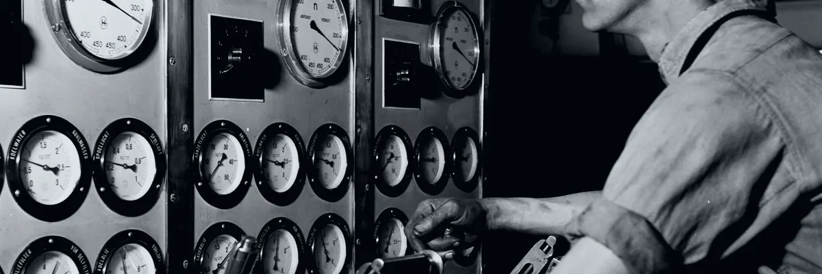 Black-and-white archive photo of a second engineer at an engine control panel full of round gauges in a ship's engine room